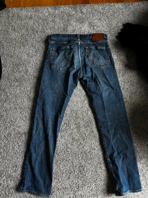 Blå raka jeans med fem fickor - Säljer ett par klassiska blå jeans med rak passform och fem fickor. Jeansen har en mörk tvätt och detaljer som nitar och läderpatch bak i midjan. Materialet är slitstark denim i bomull. Perfekta för en avslappnad och tidlös stil.