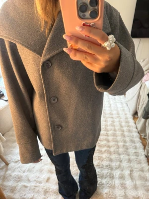 Grå rock från Gina Tricot - Snygg grå rock från Gina Tricot med bred krage och tre knappar framtill. Rocken har en rak passform och är tillverkad i ett mjukt tyg som känns skönt mot huden. Perfekt för dig som vill ha en stilren och enkel look.