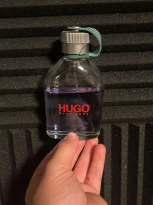 Hugo Boss parfym med lila vätska - En stilren parfymflaska från Hugo Boss med lila vätska. Flaskan är genomskinlig med rundade former och har ett silverfärgat lock med en grön tygrem. Märkets logga syns tydligt i rött på framsidan. Perfekt för dig som gillar klassisk design.