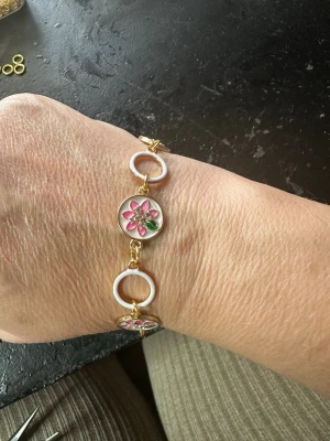 Armband med rosa blommor och gulddetaljer - Unikt armband med vita ringar och runda länkar dekorerade med rosa och gröna blommor. Guldfärgade detaljer ger en lyxig känsla och armbandet har ett magnetlås för enkel användning. Perfekt för dig som gillar färgglada accessoarer och vill sticka ut.