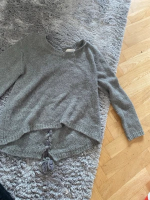 Grå stickad tröja med flätad detalj - Mysig grå stickad tröja med lång ärm och unik flätad detalj längs ryggen. Tröjan har en något längre bakdel och rundad halsringning. Perfekt för dig som gillar en avslappnad men ändå trendig look.