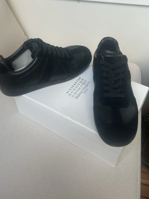 Maison Margiela  - Stilrena svarta sneakers med ovandel i både läder och mocka. Klassisk låg modell med svarta skosnören och diskret design. Perfekta för dig som gillar minimalistisk och clean stil. Sitter skönt på foten och passar till det mesta. Nästan helt nya använda 2 gånger 