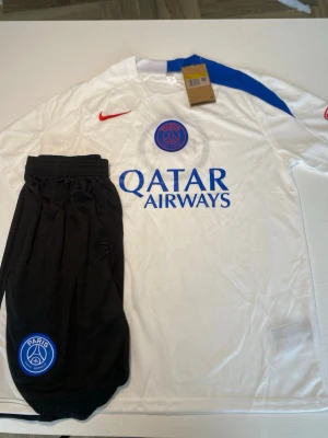 PSG bortaställ vit/blå Nike - Säljer ett PSG bortaställ från Nike med vit tröja och blå detaljer samt svarta shorts. Tröjan har PSG-logga, Qatar Airways-tryck och röd Nike-swoosh. Perfekt för fotbollsträning eller match. Materialet är lätt och andas bra.
