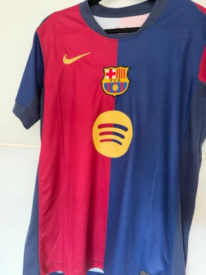 FC Barcelona matchtröja Lamine Yamal - Säljer en officiell FC Barcelona matchtröja från Nike med Lamine Yamal och nummer 19 på ryggen. Tröjan är röd och blå med klubbmärke, Nike-logga och sponsorer i gult. Tillverkad i lätt och ventilerande material, perfekt för en fet outfit eller bara till träning. Kan gå ner i pris vid snabb affär