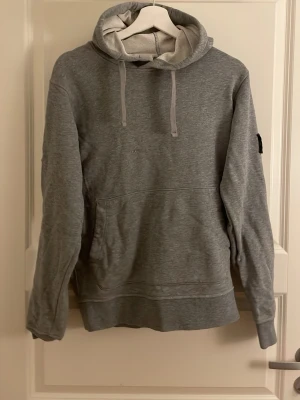 Grå hoodie med huva och magficka - Snygg grå hoodie med huva och dragsnören samt en stor magficka framtill. Tröjan har en patch på vänster ärm och är tillverkad i mjukt sweatshirtmaterial. Perfekt för en avslappnad och trendig look.
