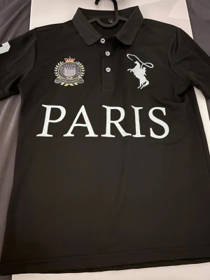 Svart pikétröja med Paris-tryck - Svart pikétröja med vit text 'PARIS' framtill, emblem på bröstet och en stor vit polospelare. Nummer 3 på ena ärmen. Klassisk krage med knappar och korta ärmar. Perfekt för en sportig och stilren look.