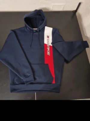 Mörkblå hoodie med tryck - Snygg mörkblå hoodie med huva och dragsnören. Framsidan har ett vertikalt tryck i vitt och rött med text och logga. Stor magficka framtill och ribbade muddar vid ärmar och nederkant. Perfekt för en avslappnad streetwear-look.