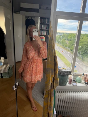 Blommig omlottklänning i rosa och orange - Kort omlottklänning med blommigt mönster i rosa och orange toner. Klänningen har långärmade, luftiga ärmar och markerad midja med resår. V-ringad framtill och volangkantad nederdel. Perfekt sommar klänning😍