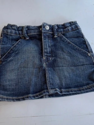 Kort jeanskjol med fickor och bälte - Säljer en blå kort jeanskjol med fram- och bakfickor, bälteshällor och dekorativa knappar. Kjolen har en klassisk denimlook med kontrastsömmar och ett litet bälte bak. Perfekt för dig som gillar en avslappnad och trendig stil. För barn. Storlek 110 