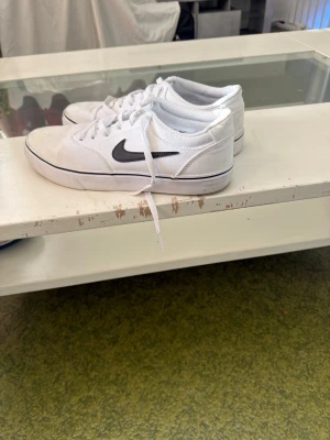 Vita Nike sneakers med svart swoosh - Säljer ett par vita Nike sneakers med klassisk svart swoosh på sidan. Skorna har lågt skaft, vita snören och en tunn svart rand runt sulan. Ovandelen är i textil och mesh för skön ventilation. Perfekta för dig som gillar stilrena och sportiga skor. Ända problemet är att det är lite smutsigt men går att få bort smutsen