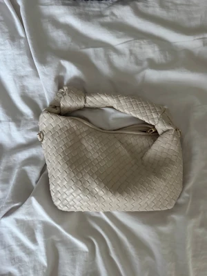 Beige flätad handväska med knut - Snygg beige handväska med flätad design och en unik knutdetalj på handtaget. Väskan har en mjuk, rundad form och stängs med dragkedja. Perfekt för dig som gillar trendiga accessoarer och stilren look.