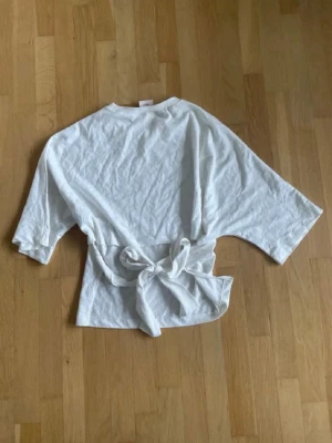 Knyt topp - Säljer en vit omlottopp med breda knytband i midjan och vida trekvartsärmar. Toppen har en avslappnad passform och är gjord i mjukt bomullstyg. Perfekt att styla med jeans eller kjol för en trendig look.