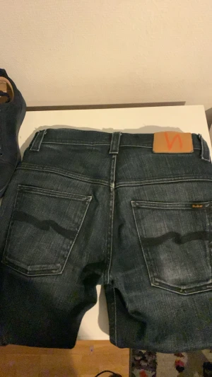 Mörkblå jeansshorts från Nudie - Säljer ett par mörkblå jeansshorts från Nudie med klassisk femficksdesign och kontrastsömmar. Shortsen har en snygg tvättad look och Nudie-loggan på bakfickan. Perfekta för dig som gillar stilrena denimshorts.