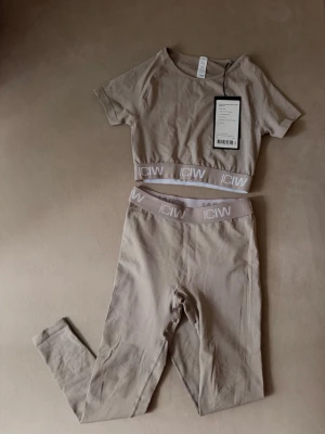Beige set från ICANIWILL XS - Snyggt beige träningsset från ICANIWILL med kortärmad crop top och höga tights. Båda delarna har elastisk midja med logga. Tröjan är aldrig använt, tights använda 2-3 gånger.