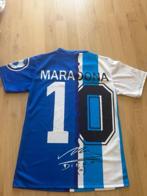 Blåvit Maradona fotbollströja - Säljer en retroinspirerad fotbollströja i blått och vitt med Maradonas namn och nummer 10 på ryggen. Tröjan har tryckta detaljer som AFA-logga, italiensk sköld, och Maradonas ansikte framtill. V-ringad hals och korta ärmar. Materialet är lätt och andas, perfekt för fotboll eller samling.
