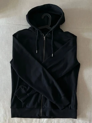 Svart zip hoodie med huva  - Säljer en stilren svart hoodie med dragkedja framtill och justerbar huva med snören. Hoodien har fickor på sidorna och ribbade muddar vid ärmslut och nederkant. Perfekt för en avslappnad och cool look. Ifrån lager 157, skriv för mer frågor!