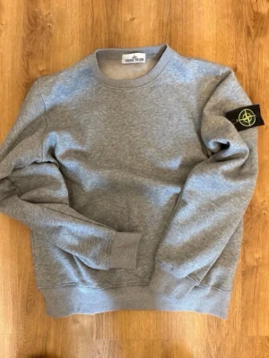 Grå sweatshirt från Stone Island - Säljer en stilren grå sweatshirt från Stone Island med klassisk rund hals och ribbade muddar. Tröjan har den ikoniska Stone Island-loggan på vänster ärm. Perfekt för dig som gillar enkel men snygg streetwear.