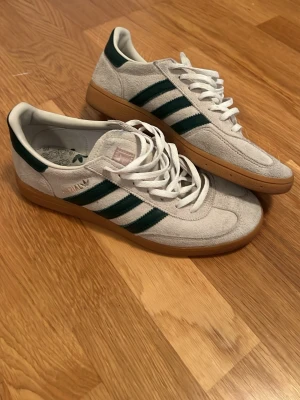 Vita Adidas spezial med gröna ränder - Snygga vita sneakers från Adidas med klassiska gröna ränder på sidorna och beige sula. Skorna har en låg profil och är gjorda i mocka med vita skosnören. Perfekta för dig som gillar retrostil och streetwear.
