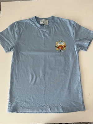 Ljusblå Casablanca t-shirt  - Säljer en ljusblå t-shirt från Casablanca med stort färgglatt tryck på ryggen och mindre tryck på bröstet. T-shirten har korta ärmar och är gjord i mjuk bomull. Perfekt för dig som gillar unika prints.