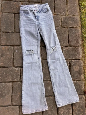 Ljusblå bootcut jeans med slitningar - Säljer ett par ljusblå bootcut jeans med slitna detaljer på båda knäna och broderade fickor bak. Jeansen har låg midja och klassisk femficksmodell. Perfekta för dig som gillar en avslappnad och trendig stil. 