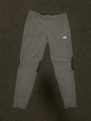 Grå träningsbyxor från The North Face - Snygga grå träningsbyxor från The North Face med svarta detaljer vid knäna och logga på låret. Byxorna har smal passform, dragkedjefickor och är tillverkade i ett stretchigt funktionsmaterial som passar perfekt för träning och utomhusaktiviteter. 🚨‼️Pris kan diskuteras vid snabb affär ‼️🚨