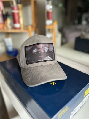 Beige keps med mesh och tryck - Säljer en beige keps med mesh baktill och ett stort svartvitt tryck framtill som föreställer två personer. Kepsen har böjd skärm och justerbar snapback. Perfekt för dig som vill ha en unik accessoar. Om du har några frågor, skriv till mig så svarar jag så snabbt jag kan