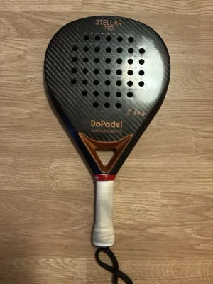 DoPadel Stellar Pro padelracket - Snygg padelracket från DoPadel, modell Stellar Pro Superior Series. Racketen är svart med bruna och vita detaljer, har rund form och många hål i träffytan. Handtaget är lindat med vitt greppband och har handledsrem. Perfekt för dig som vill ta ditt padelspel till nästa nivå.