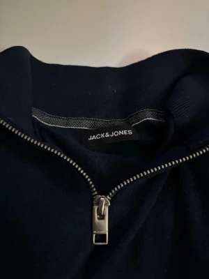 Mörkblå halvzip från Jack & Jones - Mörkblå tröja från Jack & Jones med halv dragkedja och hög krage. Enkel och stilren design utan synliga loggor på framsidan. Perfekt för lager på lager och passar till det mesta. Materialet ser ut att vara mjukt och bekvämt.