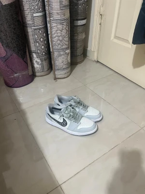 Nike Airforce  - Snygga sneakers i vitt och ljusgrått med en svart swoosh som har ett unikt mönster. Skorna har låga skaft, vita sulor och matchande grå skosnören. Perfekta för dig som vill sticka ut med en clean och modern look.