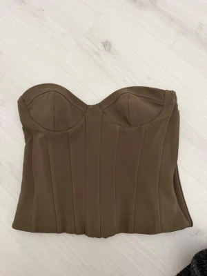 Brun korsett från ZARA - Snygg brun korsett från ZARA i storlek XS. Modellen är axelbandslös med markerade sömmar och formpressade kupor för en smickrande siluett. Tillverkad i ett mjukt och stretchigt material som sitter bekvämt mot kroppen. Perfekt att styla till olika looks.