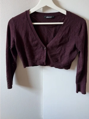 Vinröd cropped kofta från Gina Tricot - Snygg vinröd cropped kofta från Gina Tricot med v-ringning och knappar framtill. Koftan har långa ärmar och ribbade muddar vid ärmslut samt nederkant. Perfekt att styla över ett linne eller klänning för en trendig look.