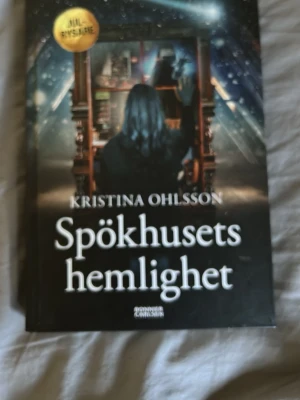 Spökhusets hemlighet av Kristina Ohlsson - En spännande och mystisk bok av Kristina Ohlsson, 'Spökhusets hemlighet', med ett mörkt omslag som visar en person framför en bokhylla i ett spöklikt rum. Perfekt för dig som gillar rysare och mysterier med vinterkänsla.
