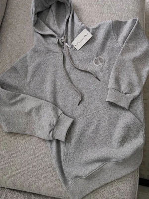 Grå hoodie från Christian Dior - Snygg grå hoodie från Christian Dior med broderad CD-logga på bröstet. Klassisk modell med huva och dragsnören samt ribbade muddar vid ärmar och nederkant. Perfekt för en avslappnad och trendig look.