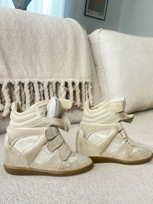 Isabel Marant, storlek 36 - Snygga beige sneakers från Isabel Marant med kilklack och kardborreband. Skorna har en mix av mocka och textil, samt gummisula med mönster. Perfekta för dig som vill ha både stil och höjd i vardagen. Köpta från plick i vintras!! 💛