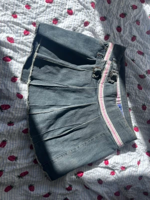 Kort jeanskjol med rosa rand - Säljer en kort jeanskjol i blå denim med rå kant och plisserad framsida. Kjolen har två dekorativa spännen och ett rosa-vitt randigt band i midjan. Perfekt för dig som gillar en sportig och trendig look.