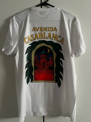 Vit Avenida Casablanca t-shirt - Vit t-shirt från Casablanca med tryck både fram och bak. På ryggen finns ett stort färgglatt motiv med texten 'Avenida Casablanca' och en byggnad i nattscen, inramad av gröna blad. Framtill finns ett mindre tryck med samma tema. Klassisk passform och rund hals.