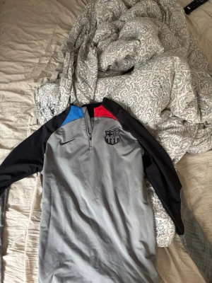 Grå Nike FC Barcelona träningströja - Snygg grå långärmad träningströja från Nike med FC Barcelona-logga på bröstet. Tröjan har svarta ärmar, blå och röda detaljer vid axlarna samt halv dragkedja framtill. Tillverkad i Dri-FIT-material som andas och passar perfekt till träning.