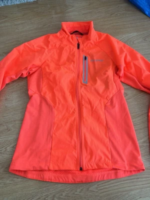 Neonorange träningsjacka med dragkedja - Snygg neonorange träningsjacka med hel dragkedja framtill och en bröstficka med dragkedja. Jackan har hög krage och är tillverkad i ett lätt, stretchigt material som passar perfekt för löpning eller utomhusträning.