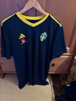 Blå Sverige fotbollströja Adidas - Mörkblå fotbollströja från Adidas med gula detaljer och Sveriges landslagsemblem på bröstet. Tröjan har korta ärmar, gul V-ringad krage och tre gula ränder på axlarna. Materialet är lätt och andas, perfekt för träning eller match.