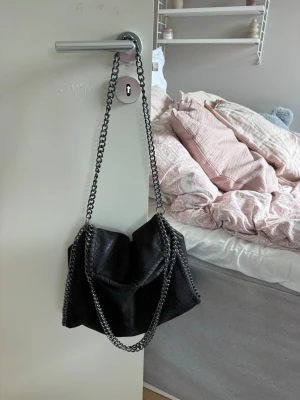 Svart handväska med kedjerem - Snygg svart handväska i mjukt mockaliknande material med silverfärgade kedjeremmar och dekorativa sömmar längs kanterna. Perfekt för dig som gillar en edgy och trendig look. Väskan har en slouchy form och känns riktigt cool till alla outfits.