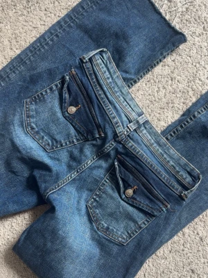 Blå bootcut jeans med fickdetaljer - Säljer ett par blå bootcut jeans med snygga fickdetaljer och knappar bak. Stretchigt material i storlek 38 från H&M, nytt skick🌸