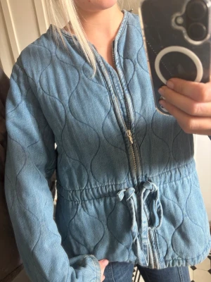 Quiltad jeansjacka med dragkedja - Snygg quiltad jeansjacka i ljusblå denim med vågigt mönster. Jackan har dragkedja framtill, justerbar midja med knytband och långa ärmar. Perfekt för dig som vill ha en avslappnad men trendig look.