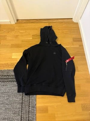 Svart hoodie med röd detalj - Säljer en svart hoodie med luva och en unik röd dragkedjedetalj på ena ärmen. Hoodien har en klassisk passform och ribbade muddar vid ärmslut och nederkant. Perfekt för dig som gillar streetwear och vill sticka ut lite extra.
