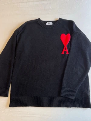 AMI Paris - Snygg svart stickad tröja från Ami med ikoniskt rött hjärta och bokstaven A broderat på bröstet. Tröjan har rund hals, ribbade muddar och är gjord i mjukt material som känns lyxigt. Perfekt för dig som gillar stilrena plagg med en twist.