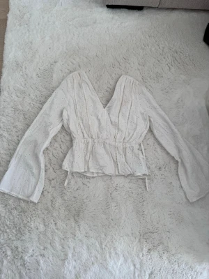 Vit omlottopp med volang och knyt - Söt vit omlottopp med långa ärmar och volangkant nedtill. Toppen har knyt i midjan och v-ringad hals. Materialet är lätt och har broderade detaljer som ger en romantisk känsla. Perfekt för dig som gillar en feminin och trendig stil.