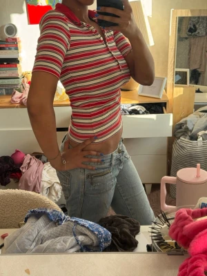Randig ribbad topp med krage - Säljer en ribbad topp med röda, vita och svarta ränder. Modellen har korta ärmar, krage och knappar framtill. Sitter tight och har en croppad passform som ger en retrovibe. Perfekt till jeans eller kjol.