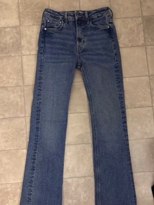 Blå flare jeans med rå kant - Säljer ett par klassiska blå jeans med rak passform och rå, fransig kant längst ner på benen. Jeansen har fem fickor och stängs med knapp och dragkedja. Materialet är kraftig denim i 100% bomull. Perfekta för en avslappnad och trendig look.