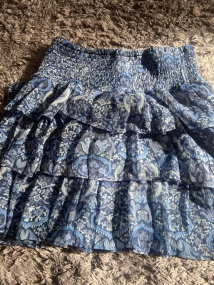 Blå volangkjol med mönster - Kort blå kjol med flera volanger och ett vackert blommigt mönster i olika blå toner. Kjolen har bred smock i midjan för skön passform och är gjord i ett lätt, luftigt material som passar perfekt till varmare dagar speciellt. Väldigt bra skick, är i storlek 164 men är naturligt lång så jag skulle säga att den kan passa på någon upp till XS/S