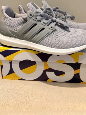 Grå Adidas Ultraboost sneakers - Snygga grå Adidas Ultraboost sneakers med stickad ovandel och klassiska tre ränder på sidan. Skorna har en vit Boost-sula för extra komfort och grå snörning. Perfekta för dig som gillar stilrena och sportiga sneakers.