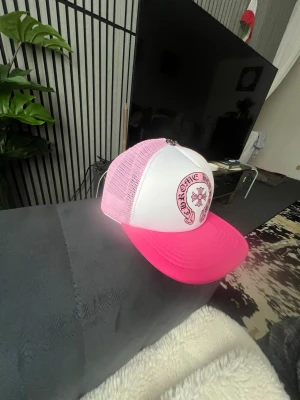 Vit och rosa truckerkeps - Säljer en snygg truckerkeps med vit front och rosa mesh samt skärm. Kepsen har ett tryck framtill med text och motiv i rosa. Justerbar baktill för perfekt passform. Perfekt accessoar för dig som vill sticka ut med färg.